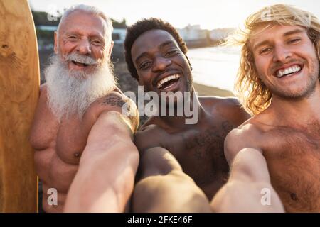 Felices surfistas en forma con diferentes edades y carrera tomando selfie mientras se divierte practicando surf juntos al atardecer Foto de stock
