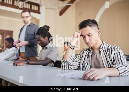 Retrato de los estudiantes que toman el examen en fila mientras están sentados en el escritorio en el auditorio, foco en el hombre asiático joven que piensa en primer plano, espacio de copia