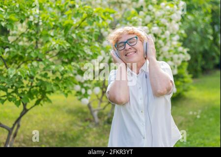 Una mujer anciana contenta disfruta de la música y sostiene los auriculares al aire libre. La anciana caucásica sonriente camina en un parque floreciente y escucha Foto de stock