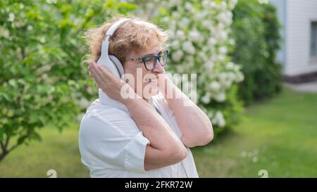 Una mujer anciana contenta disfruta de la música y sostiene los auriculares al aire libre. La anciana caucásica sonriente camina en un parque floreciente y escucha Foto de stock