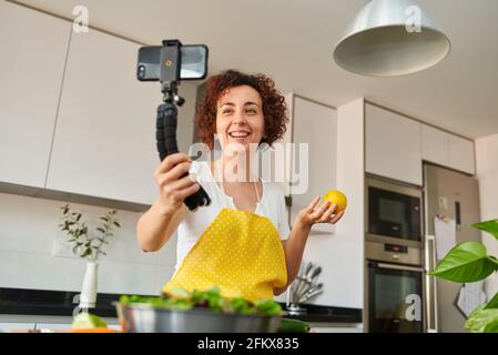Mujer youtuber se registra con su smartphone en su cocina mientras prepara una receta de ensalada, hay luz natural y ella está usando un amarillo A Foto de stock