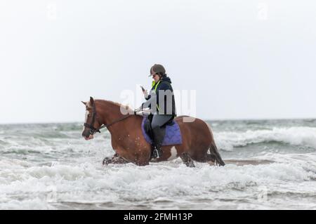 Irvine, Escocia, Reino Unido. 9th de mayo de 2021. Clima en el Reino Unido: Un jinete que usa un teléfono móvil a caballo en las olas en Irvine Beach. Crédito: Skully/Alamy Live News Foto de stock