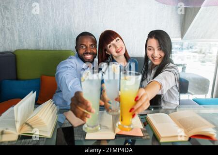 Grupo de tres amigos multirraciales felices trachando vasos con coloridos cócteles de jugo en la cafetería. Jóvenes alegres y atractivos que pasan tiempo en la cafetería, leyendo libros y bebiendo jugo