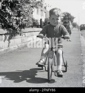 1950s, histórico, un niño pequeño en un pavimento de la calle que monta su triciclo, un juguete del pedal del tri-ang, Inglaterra, Reino Unido. Fundada en 1919, Tri-ang Toys fue el nombre de marca de la compañía británica Lines Brothers, que creó tantos negocios, que en algún momento fue la compañía de juguetes más grande del mundo. Se plegaron en 1971. Foto de stock