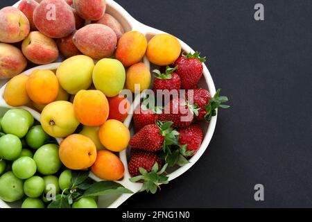 Vista superior de frutas frescas de verano, ciruela, melocotón, albaricoque, fresa en plato sobre fondo negro, espacio de copia, concepto de comida. Foto de stock