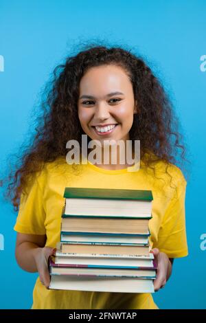 Estudiante niña tiene un montón de libros universitarios de la biblioteca sobre fondo azul en el estudio. La mujer sonríe, ella está feliz de graduarse. Foto de stock