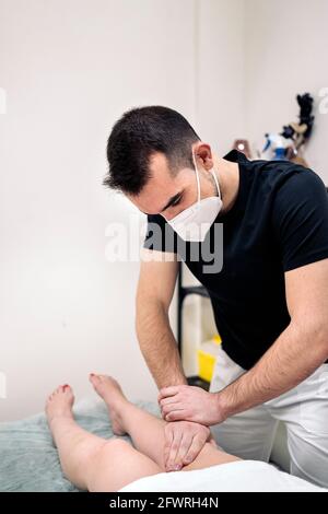 Fisioterapeuta masculino con máscara facial que da masaje de piernas a una mujer no reconocida que se encuentra en la camilla. Foto de stock