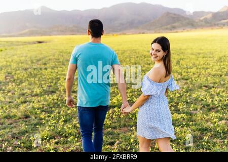 Mujer sonriente sosteniendo la mano del hombre mientras caminaba en el prado Foto de stock