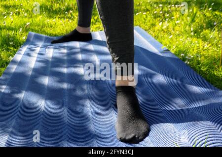 Mujer con leggings grises y calcetines negros, practicando yoga pie en el guerrero dos posición una alfombra azul en el césped verde al aire libre de stock -