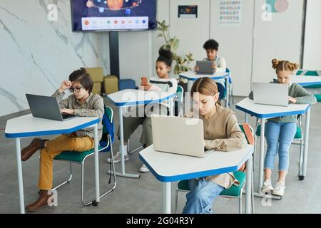 Un grupo diverso de niños pequeños que usan computadoras portátiles mientras están sentados en los escritorios en el salón de clases de la escuela, espacio para copiar