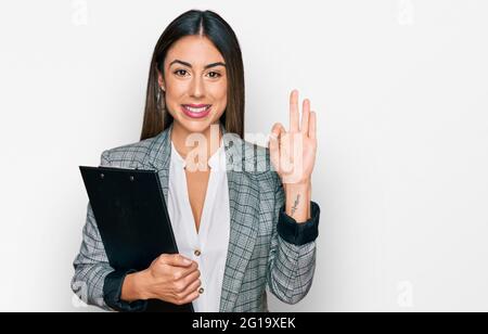 Joven mujer hispana que lleva ropa de negocios sosteniendo el portapapeles haciendo el signo correcto con los dedos, sonriendo amable gesturing excelente símbolo Foto de stock