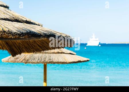 (Enfoque selectivo) Vista impresionante de una sombrilla en primer plano y un yate de lujo navegando en un hermoso mar turquesa en el fondo. Foto de stock