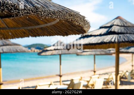 (Enfoque selectivo) Vista impresionante de una sombrilla en primer plano y sillas de sol borrosas en una playa bañada por un hermoso mar turquesa. Foto de stock