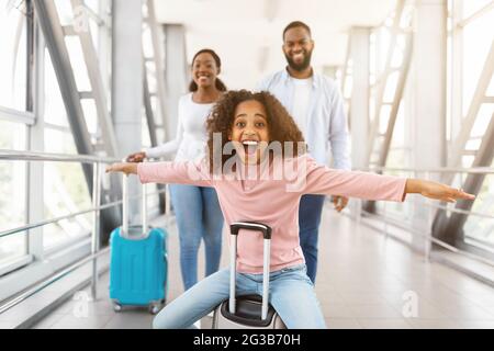 Por fin unas vacaciones. Retrato de la alegre muchacha afro-americana emocionada sentada en el equipaje y riendo, extendiendo los brazos fingiendo volar, imitando aire pl Foto de stock