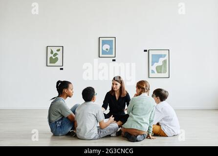 Vista completa en un grupo diverso de niños sentados en círculo durante la clase en la galería de arte, espacio de copia