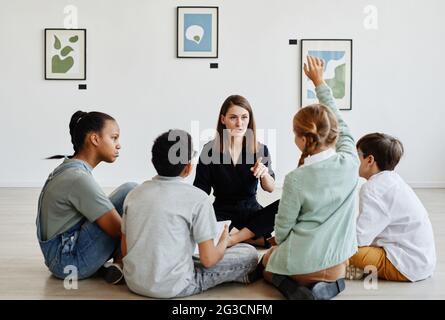 Vista completa a un grupo diverso de niños sentados en círculo durante la clase en la galería de arte con maestra