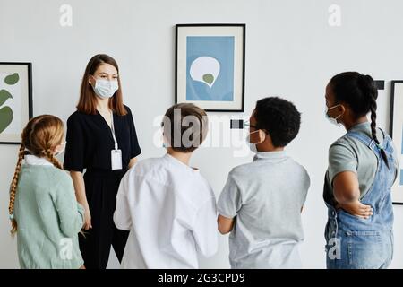 Vista posterior a un grupo diverso de niños mirando pinturas en la galería de arte con una maestra con máscara