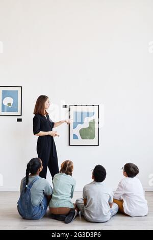 Vista vertical de la espalda a un grupo diverso de niños sentados en el piso en la galería de arte moderno y mirando las pinturas, el espacio de copia