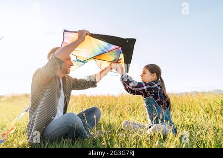 Sonriente niña sentada en el césped con el padre ayudándola a preparar colorido juguete arcoiris cometa para volar. Momentos felices de la infancia en familia o al aire libre t Foto de stock