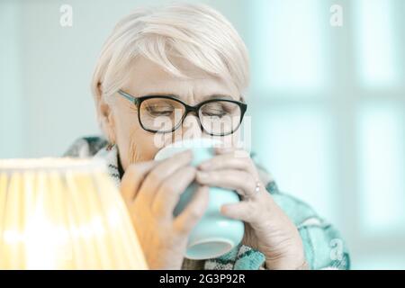La mujer mayor envuelta en un cuadros de cuadros de cuadros es beber té en el hogar Foto de stock