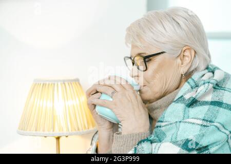 La mujer mayor envuelta en un cuadros de cuadros de cuadros es beber té en el hogar Foto de stock