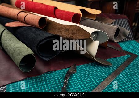 Enrollado muestras de texturas de cuero auténtico de diferentes colores sobre un fondo de cuero, sobre una mesa con alfombrillas y herramientas para cortar piezas de lea Foto de stock