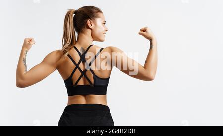Deporte y mujeres. Vista posterior de atleta de fitness fuerte, culturista femenino, flexionando los músculos, mostrando el cuerpo en forma, bíceps y espalda atlética, sonriendo
