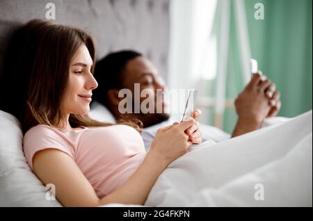 Concepto de adicción a gadgets. Familia multiétnica joven que utiliza smartphones mientras está tumbado en la cama, copia espacio