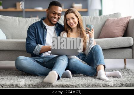 Pasatiempo doméstico. Jóvenes y alegres cónyuges interraciales descansando con el ordenador portátil en casa