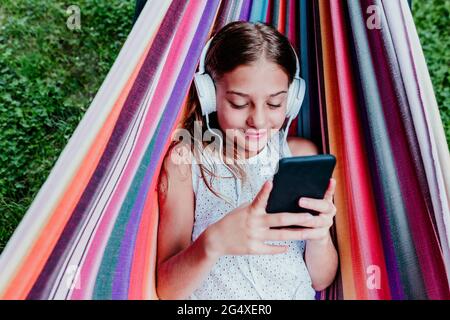 Chica sonriente usando auriculares usando el teléfono móvil mientras descansa en una hamaca Foto de stock