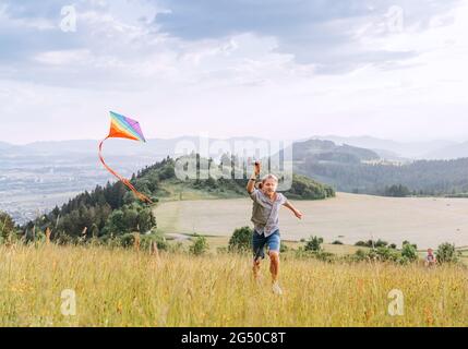 Chico adolescente sonriente con cometa de colores volando en el prado de hierba alta en los campos de montaña. Momentos felices de la infancia o tiempo al aire libre pasando conce Foto de stock