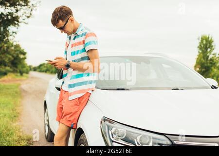 Hombre joven guapo en ropa de verano y gafas de sol de pie cerca de su coche blanco de lujo y utilizando smartphone, sosteniendo en la mano, al aire libre en un sol de verano