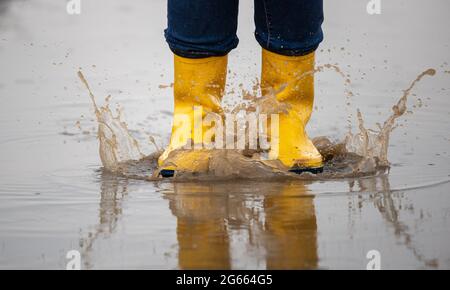 Primer plano de las piernas femeninas en botas de agua amarillas saltando en charcos después de la lluvia en el campo Foto de stock