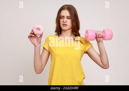 Hermosa mujer de edad joven con cuerpo delgado que sostiene en manos rosa mancuerna y donut, mirando pastel con expresión facial repugnante. Estudio interior Foto de stock