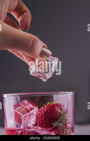 La mano femenina de primer plano pone el cubo de hielo en un vaso de fresas. Foto de stock