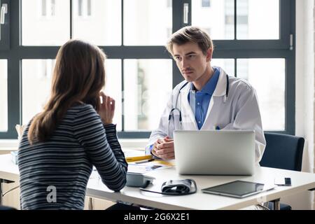 Devoto joven doctor sosteniendo una placa de rayos X mientras escucha con atención a su paciente durante una consulta privada en la oficina Foto de stock