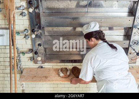 Una mujer panadero sacando panes del horno de panadería poniéndolos en una tabla Foto de stock