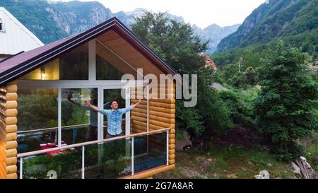 Un hombre joven y guapo está en el balcón de un apartamento de campo que mira con una sonrisa en su cara sobre un fondo de montañas. Descanse en el Foto de stock