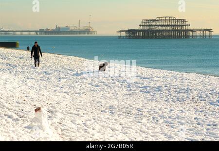 Nieve en la playa, Brighton, Inglaterra, con el Brighton Palace Pier y restos del West Pier detrás. Hombre perro caminante en la nieve y muñeco de nieve en primer plano. Foto de stock