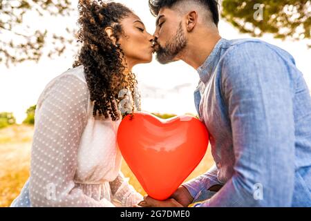  Fotografía de perfil de pareja multirracial en amor besando al atardecer en la naturaleza con efecto retroiluminado por el sol en globo rojo en forma de corazón entre ellos. Romántico Fotografía de