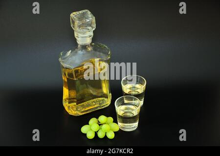 Grappa, alcohol fuerte en una botella y dos vasos, al lado de un manojo de uvas verdes sobre fondo negro. Primer plano. Foto de stock