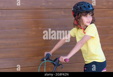 niño con pelo largo y casco de seguridad se divierte montando una bicicleta. fondo de pared de madera. espacio de copia Foto de stock
