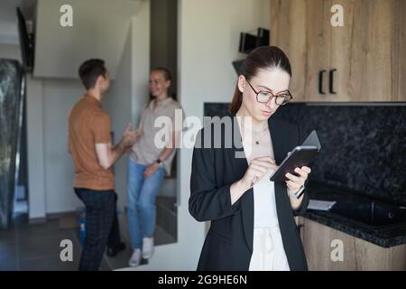 Cintura para arriba retrato de agente inmobiliario femenino usando smartphone con pareja joven en el fondo, espacio de copia