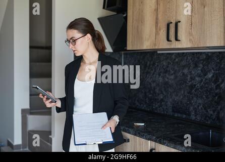 Retrato gráfico de agente inmobiliario femenino con smartphone mientras se encuentra junto a la cocina en casa para la venta, copia espacio
