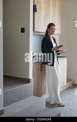 Retrato vertical de largo completo de agente inmobiliario femenino con smartphone mientras se encuentra junto a la cocina en casa para la venta, espacio de copia