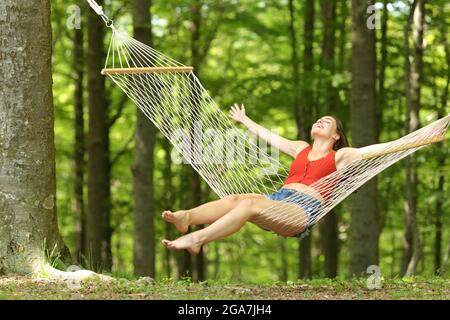 Mujer emocionada balanceándose en una hamaca celebrando vacaciones en un hermoso bosque verde Foto de stock