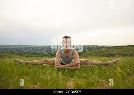 Instructor de yoga masculino serio que practica el ejercicio de estiramiento de la pierna en prado verde del verano