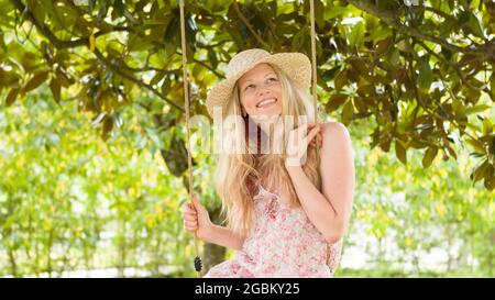 Mujer rubia sonriente con sombrero de paja, balanceándose en el columpio en el jardín de la casa, el concepto de vacaciones de ocio y verano Foto de stock