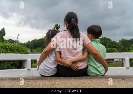 Vista posterior de la madre asiática amorosa abrazar a sus hijos sentados en el puente, la madre negra cariñosa abrazar a su hijo, relajándose mirando a la nube negra, el padre conforta a su hijo