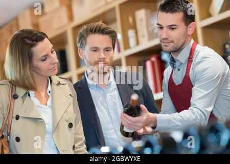 hombre vendedor y pareja con botellas de vino de pie en el estante